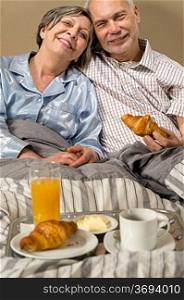 Happy retired couple eating croissant for breakfast in bed