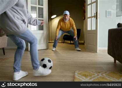 Happy retired couple playing football in home living room. Elderly husband trying to catch ball while older wife starting to kick. Fun entertainment on weekend. Happy retired couple playing football in home living room