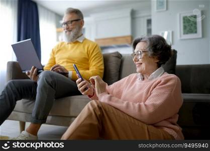 Happy retired couple using digital gadget while resting in living room at home. Elderly wife chatting friends on smartphone and senior husband messaging social media on laptop computer. Happy retired couple using digital gadget while resting at home