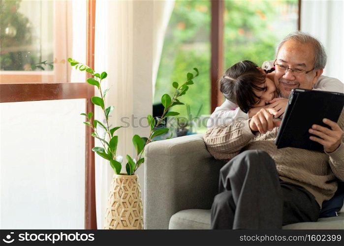Happy retirement elderly man sitting on sofa at living room with granddaughter using digital tablet together. Multigenerational family with technology concept.