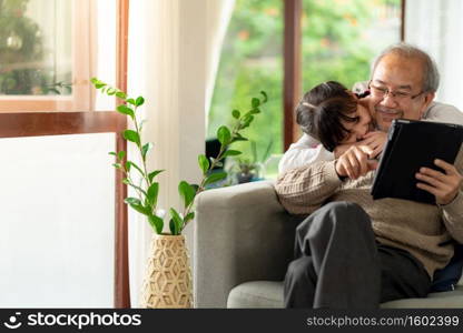 Happy retirement elderly man sitting on sofa at living room with granddaughter using digital tablet together. Multigenerational family with technology concept.