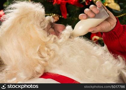 Happy Santa Claus drinking milk from glass bottle against Christmas Tree at home.