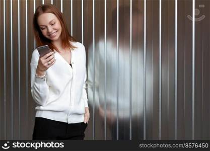 Happy satisfied browses website on cell phone, wears white jumper and black trousers, smiles happily, enjoys online communication, being successful business worker. Networking and technology