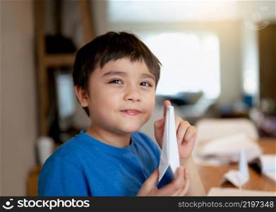 Happy school boy looking at camera with smiling face while showing origami Swan paper. Kid learning paper art origami lesson, Child having fun doing Art and Craft at home, Home schooling concept