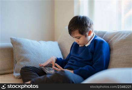 Happy schoolboy playing game online with friend on tablet,Kid using internet sending homework to the teacher,Positive child sitting on sofa relaxing alone in living room in the morning