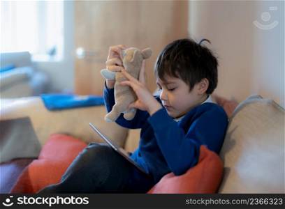 Happy schoolboy playing with dog toy and game online with friend on tablet,Kid using internet sending homework to the teacher,Positive child sitting on sofa relaxing alone in living room in the morning