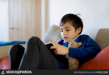 Happy schoolboy playing with dog toy and game online with friend on tablet,Kid using internet sending homework to the teacher, Positive child sitting on sofa relaxing in living roomafter school
