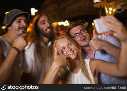 Happy selfie of friends at night. Group of happy and excited young people having good time outdoor at night. Focus on man and woman showing thumbs-up and making mobile phone selfie