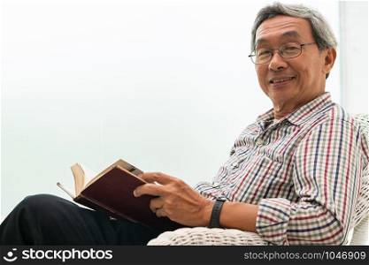 Happy senior Asian man reading book on the chair in living room at home. Retirement lifestyle and aging society concept.