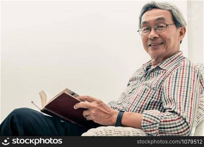 Happy senior Asian man reading book on the chair in living room at home. Retirement lifestyle and aging society concept.