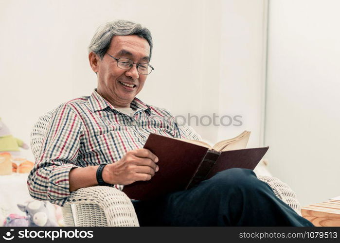 Happy senior Asian man reading book on the chair in living room at home. Retirement lifestyle and aging society concept.