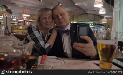 Happy senior couple taking mobile selfie during the dinner in restaurant and looking at photos. Man wearing US flag badge
