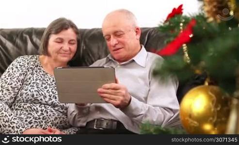 Happy senior couple using digital tablet at Christmas tree. Close up. They are out focus and then comes in focus when camera close to them.