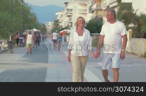 Happy senior couple walking along sidewalk on resort, man spinning a woman and excited couple continuing the way. Hotels and people in the street in background