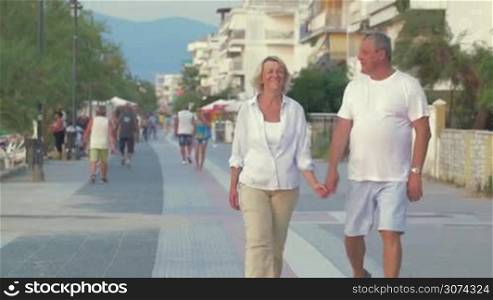 Happy senior couple walking along sidewalk on resort, man spinning a woman and excited couple continuing the way. Hotels and people in the street in background