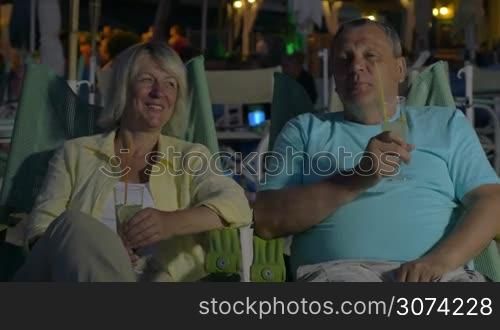 Happy senior family couple spending evening on the beach. They sitting in chaise-longues, having drinks and enjoying summer vacation