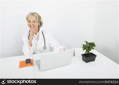 Happy senior female doctor with laptop at desk in clinic