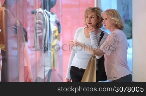 Happy senior female friends with shopping bags standing near shop window display and looking at clothes in shopping mall. Blurred boutique window with dressed mannequins on background. Shopaholic women pointing at dresses in store front.