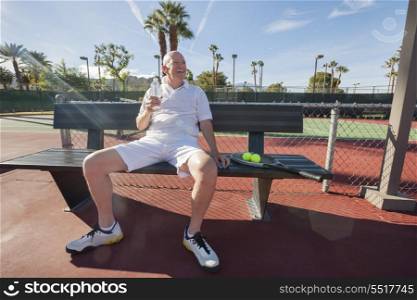 Happy senior male tennis player with water bottle sitting on bench at court