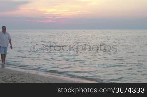 Happy senior man and walking barefoot in the shore water at sunset. Loving couple holding hands and enjoying summer time