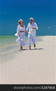 Happy senior man and woman couple running, laughing and holding hands on a deserted tropical beach with bright clear blue sky