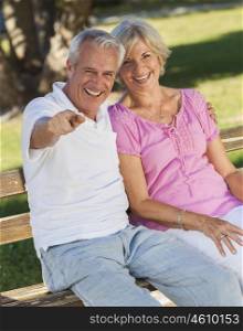 Happy senior man and woman couple sitting together laughing and pointing on a park bench outside in sunshine