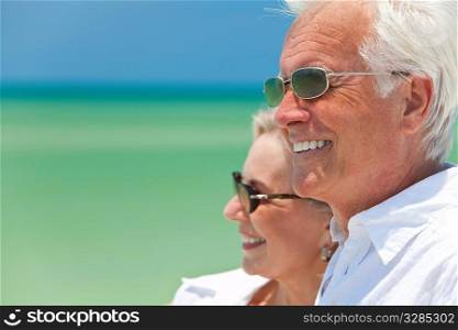 Happy senior man and woman couple together looking out to sea on a deserted tropical beach with bright clear blue sky