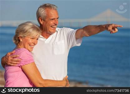Happy senior man and woman couple walking and embracing on a deserted tropical beach with bright clear blue sky, the man pointing to the horizon