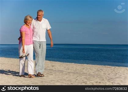 Happy senior man and woman couple walking laughing on vacation on a deserted tropical beach with bright clear blue sky and calm sea