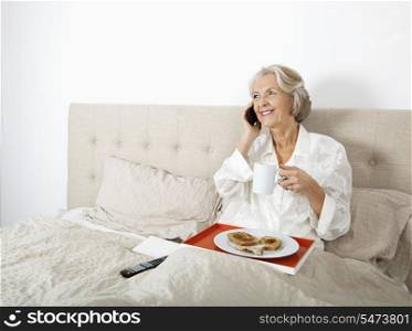 Happy senior woman answering cell phone while having breakfast in bed