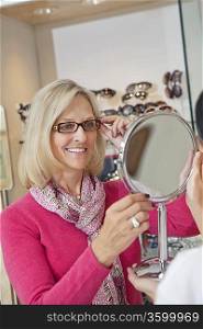 Happy senior woman trying on glasses while optician holding hand mirror