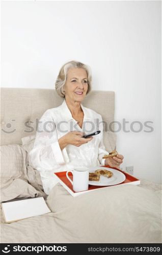 Happy senior woman watching TV while having breakfast in bed
