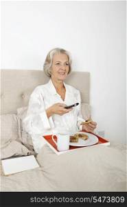 Happy senior woman watching TV while having breakfast in bed