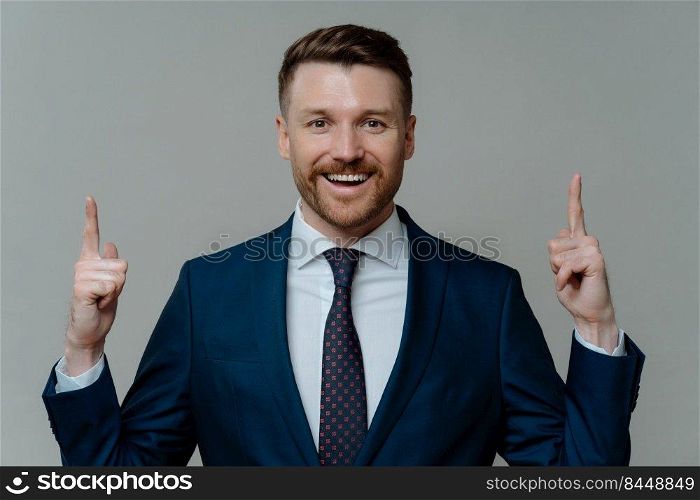 Happy sitisfied businessman in formal wear having great idea, pointing up with two forefingers and smiling at camera while standing against grey background, cheerful entrepreneur getting solution. Pleased businessman in suit pointing up with fingers at copy space