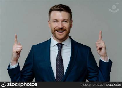 Happy sitisfied businessman in formal wear having great idea, pointing up with two forefingers and smiling at camera while standing against grey background, cheerful entrepreneur getting solution. Pleased businessman in suit pointing up with fingers at copy space