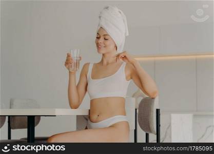 Happy slim young woman sitting on chair in kitchen with glass of pure water in her hand, relaxing after morning refreshing shower and starting new day with healthy habits. Dieting concept. Happy slim young woman sitting on chair in kitchen with glass of pure water in her hand