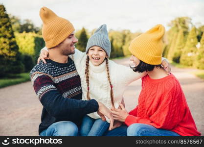 Happy small girl embrace her mother and father, have fun together, walk in beautiful park or on road. Parents look at her lovely daughter, try to do best for her. Happy family have good rest outdoors