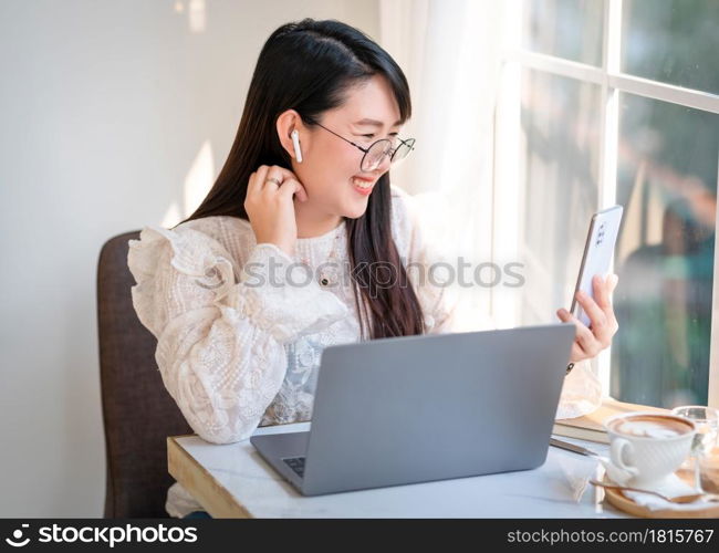 Happy smiley face asian freelance people Businesswoman wearing wireless earphones using smartphone video call camera and talking while casual working with laptop computer at the cafe,Lifestyle