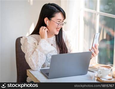 Happy smiley face asian freelance people Businesswoman wearing wireless earphones using smartphone video call camera and talking while casual working with laptop computer at the cafe,Lifestyle