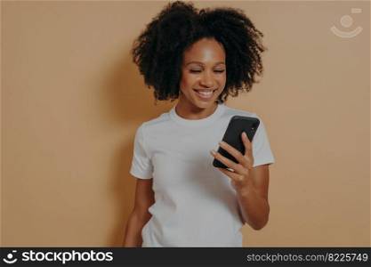Happy smiling african female student using mobile phone and smiling, reading positive news in internet or chatting with boyfriend online, holding modern smartphone while posing against sand color wall. Happy african female student using mobile phone while standing isolated over sand color background