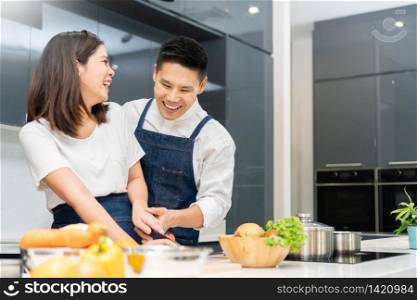 Happy Smiling Asian couple cooking in the kitchen at home. Young Man and beautiful woman in white clothes having fun preparing vegetable for salad.