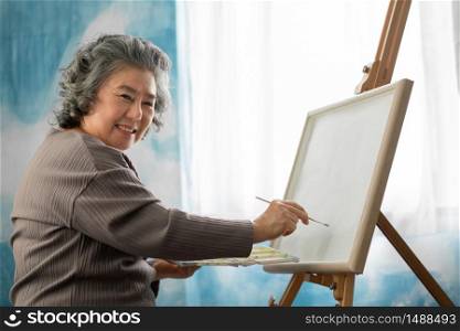 Happy smiling Asian Elderly woman oil painting on canvas at house on holidays. Artist Senior female woman in casual sweater while holding paintbrush and color palette while study art. Watercolor and Acrylic colour. Looking at camera. Retirement.