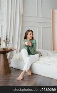Happy smiling beautiful European woman enjoys the morning and inhales the aroma of a cup of coffee in her bedroom. Enjoying free and relaxing time, the concept of a lazy weekend.