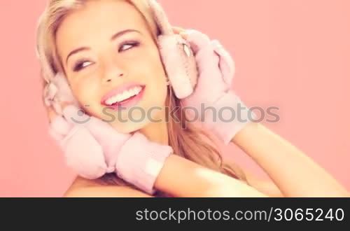 Happy smiling blonde woman in pink winter mittens and earmuffs on pink studio background