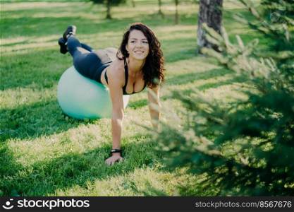 Happy smiling brunette woman has yoga exercises with fitness ball poses at park with green grass, has cheerful expression, stays fit and healthy, leads active lifestyle, has morning gymnastic