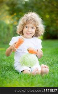 Happy smiling child with vegetables sitting on green grass in spring park. Healthy lifestyles concept