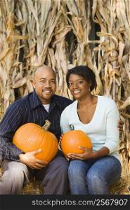 Happy smiling couple sitting on hay bales and holding pumpkins at outdoor market.
