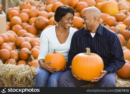 Happy smiling couple sitting on hay bales and holding pumpkins at outdoor market.