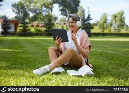 Happy smiling elderly woman talking with friends or having education webinar with teacher using videocall app on mobile digital tablet while resting in public park. Elderly woman talking with relatives using videocall app on mobile tablet