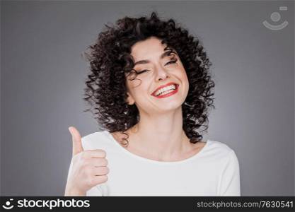 Happy smiling excited woman giving thumb up. Caucasian female with curly hair anf toothy smile on gray background. Excited woman giving thumb up